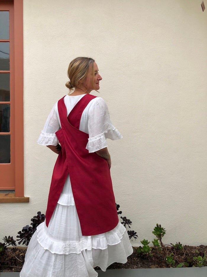cross back pinafore apron in red linen. woman is wearing it with a long white dress and standing with her back to the camera.