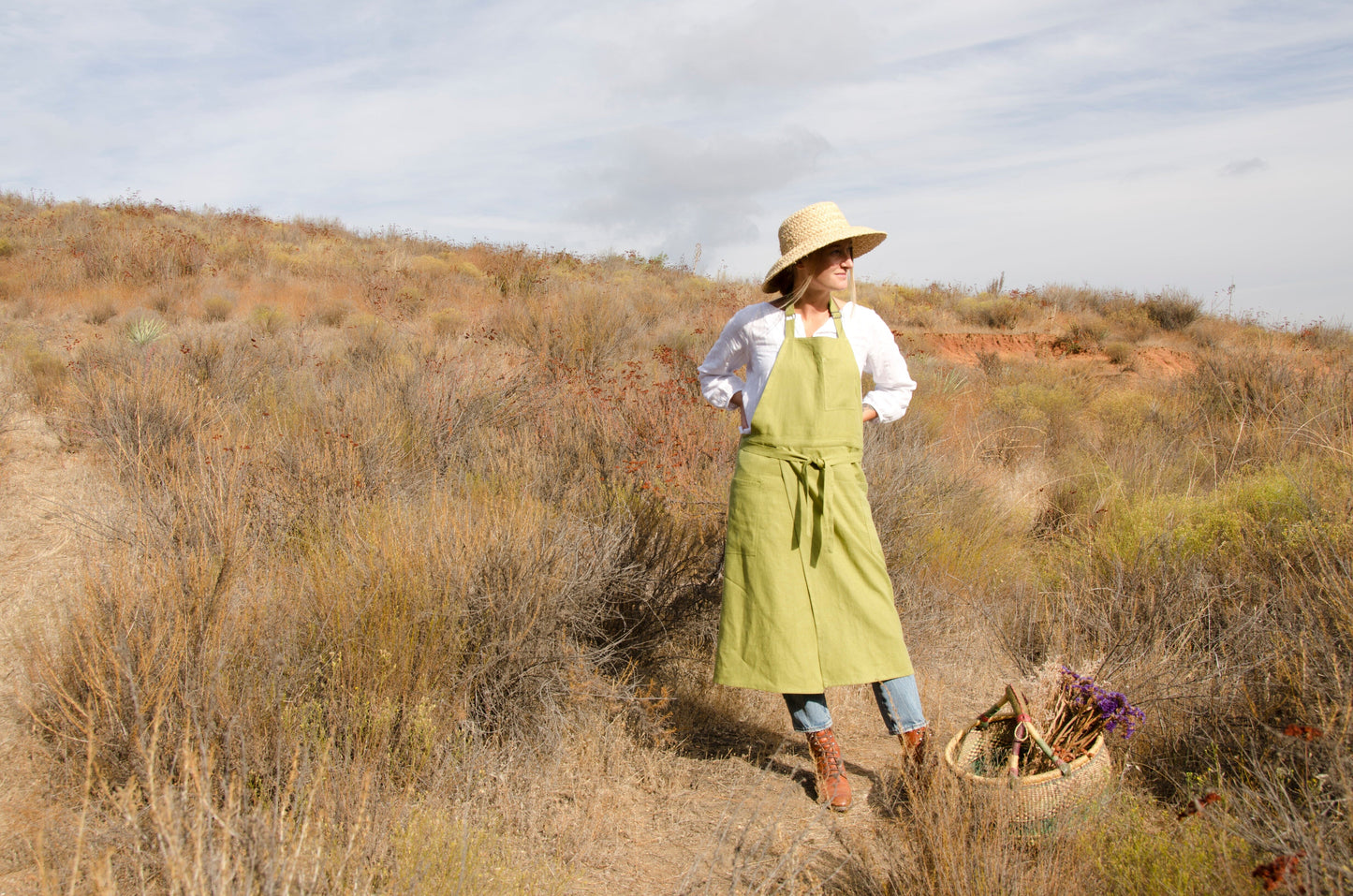 person in a field with hands on hips. wearing a white blouse, straw hat, jeans, and long green linen apron.