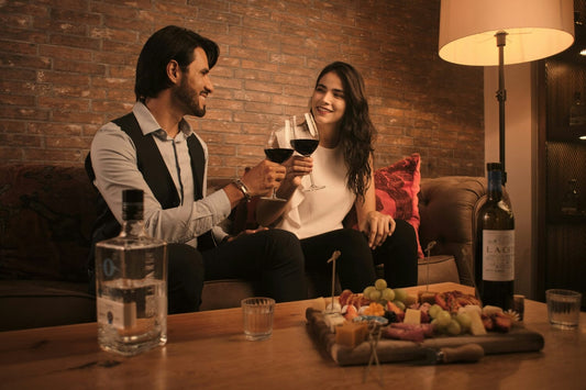 A couple in a brick studio on a couch clinking wine glasses, with wine, a bottle of tequila, and a charcuterie board on a coffee table in front of them