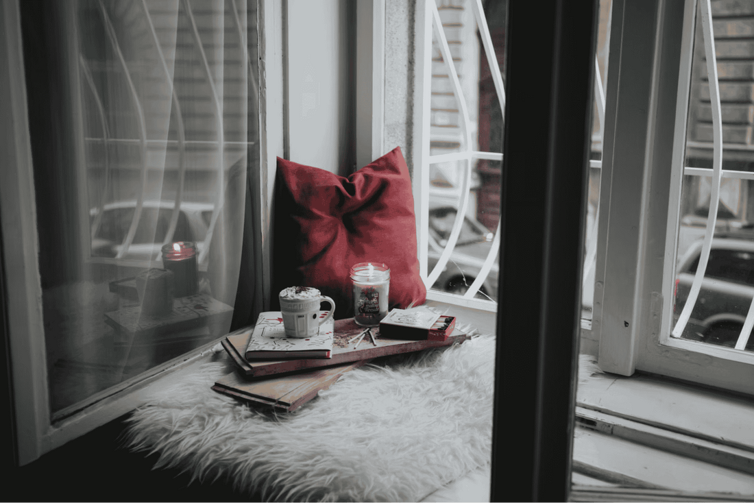 A close-up of a windowsill seat with books, a candle, a cozy rug and pillow, and a hot chocolate