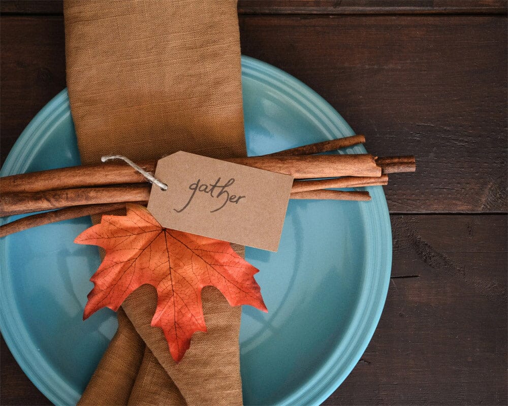 A simple fall plate setting with the word “gather” written on brown paper tied to cinnamon sticks, on top of a brown napkin and autumn leaf