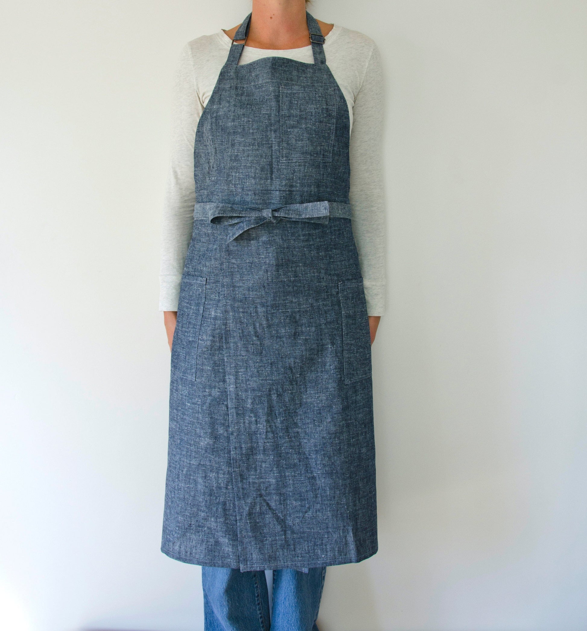 person modeling a split leg apron in linen. they are wearing a white shirt and jeans and standing in front of a white wall. the apron is denim.