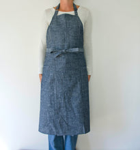 Load image into Gallery viewer, person modeling a split leg apron in linen. they are wearing a white shirt and jeans and standing in front of a white wall. the apron is denim.
