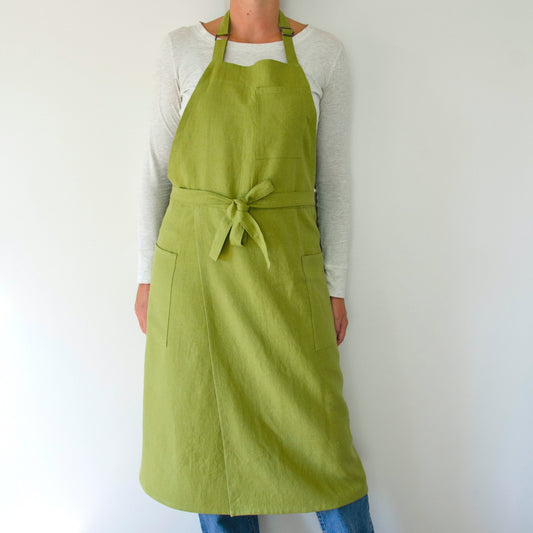 person modeling a split leg apron in linen. they are wearing a white shirt and jeans and standing in front of a white wall. the apron is green.