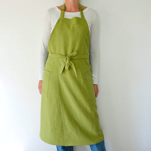 person modeling a split leg apron in linen. they are wearing a white shirt and jeans and standing in front of a white wall. the apron is green.