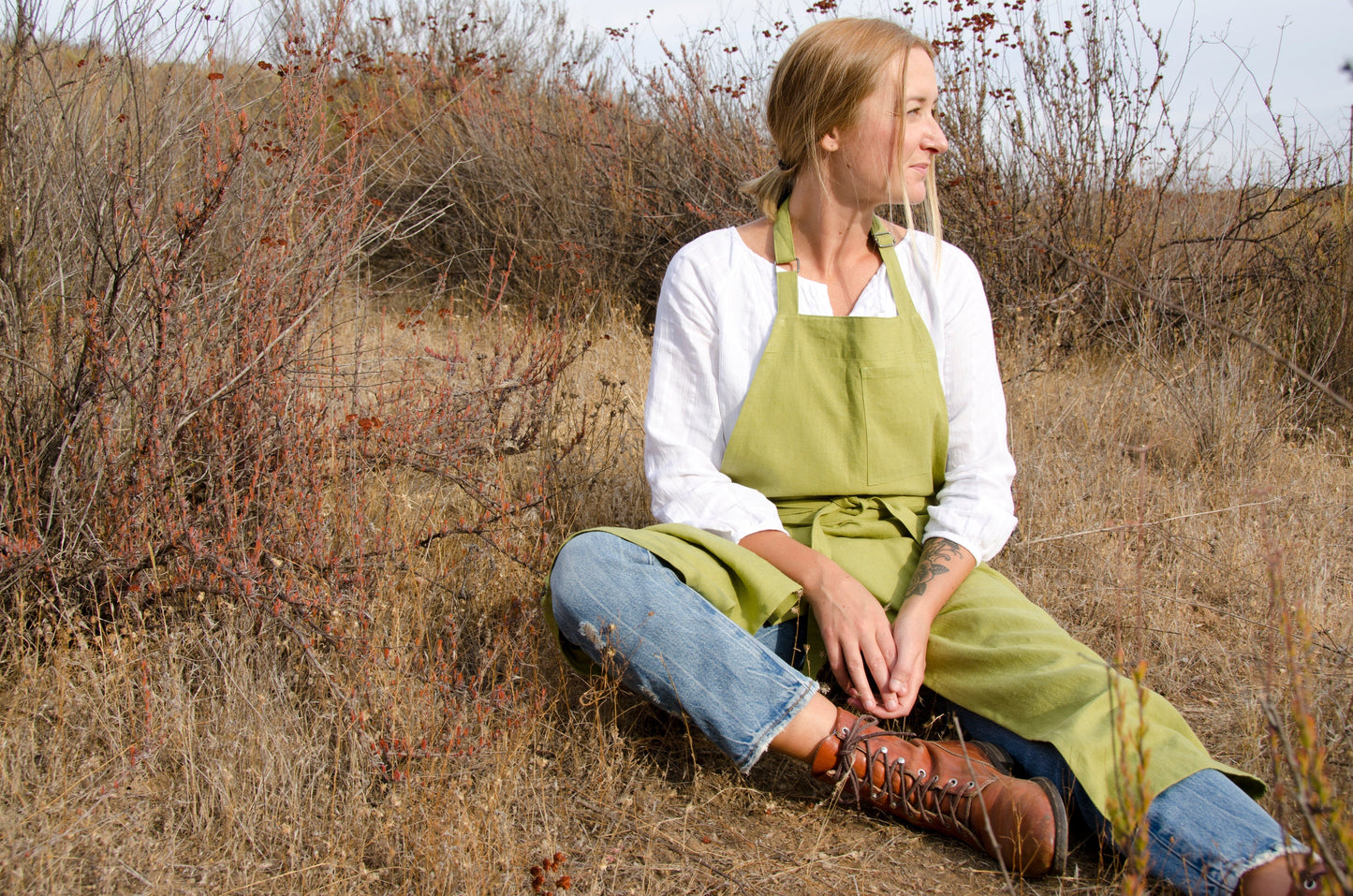 close up of a woman sitting on the ground in a dry field. she wears a white blouse, jeans, boots, and a green linen apron. 