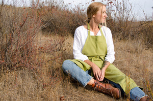 close up of a woman sitting on the ground in a dry field. she wears a white blouse, jeans, boots, and a green linen apron. 