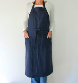 person modeling a split leg apron in linen. they are wearing a white shirt and jeans and standing in front of a white wall. the apron is blue.